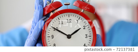 Doctor in gloves holds red alarm clock closeup. Doctor in gloves holds red alarm clock closeup. 72137381