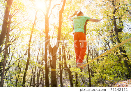 An adult bearded man balancing on a tight slackline is engaged in balancing act in the autumn forest 72142151