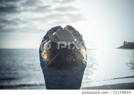 Blue color image with back view of trendy tourist enjoying the sunset at the beach with ocean and sky view - woman viewed by rear with fashion hat in outdoor leisure activity on vacation Blue color image with back view of trendy tourist enjoying the sunset at the beach with ocean and sky view - woman viewed by rear with fashion hat in outdoor leisure activity on vacation 72159644