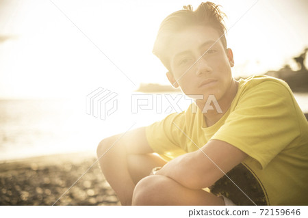 Handsome young boy portrait - caucasian male teenager age look at the camera with sunset in backlight and beach with ocean - yellow and golden colors and people outdoor concept 72159646
