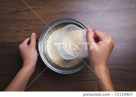 Woman hand is pouring jasmine rice into pot. Using measuring glass. on wooden table. Woman hand is pouring jasmine rice into pot. Using measuring glass. on wooden table. 72159907
