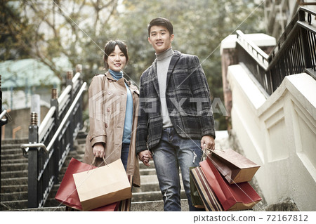 outdoor portrait of happy young asian couple standing on steps with paper shopping bags in hand 72167812