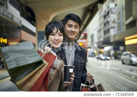 happy young asian couple waiting for bus in modern city with shopping bags in hand 72168569