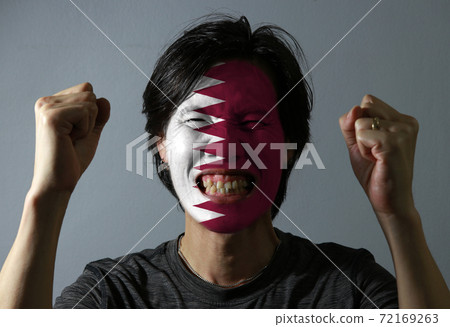 Cheerful portrait of a man with the flag of the Qatar painted on his face on grey background. 72169263
