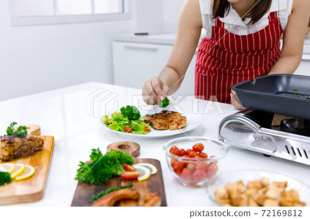 Woman chef in red apron putting parsley on pork steak with table full of food. Woman chef in red apron putting parsley on pork steak with table full of food. 72169812