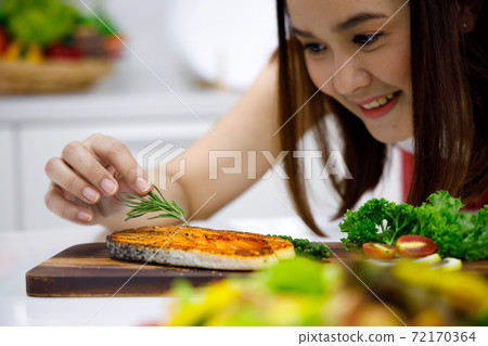 Asian woman chef garnish grilled salmon steak with rosemary. Concept woman cooking at home. Asian woman chef garnish grilled salmon steak with rosemary. Concept woman cooking at home. 72170364