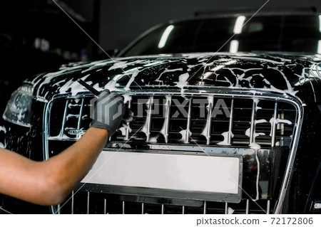 Car detailing wash. Manual cleaning with soap at car wash. Cropped image of hand of male worker in rubber glove washing car radiator grille of luxury black car with special brush and cleansing foam 72172806