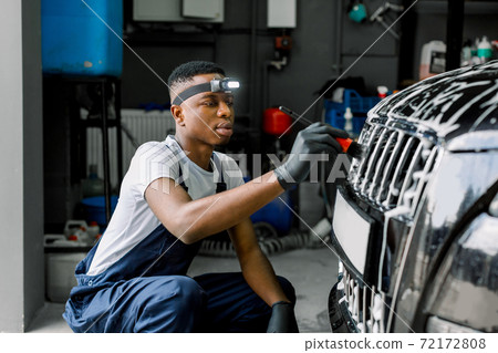Detailing manual cleaning with soap at car wash. Young black male worker in rubber gloves washes car radiator grille of luxury black car with special brush and soap. Car detailing wash. 72172808
