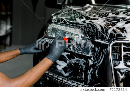Close up photo of hands of auto service male worker in black protective gloves cleaning car hood and headlights with foam and soft brush. Carwash and detailing. Close up photo of hands of auto service male worker in black protective gloves cleaning car hood and headlights with foam and soft brush. Carwash and detailing. 72172814