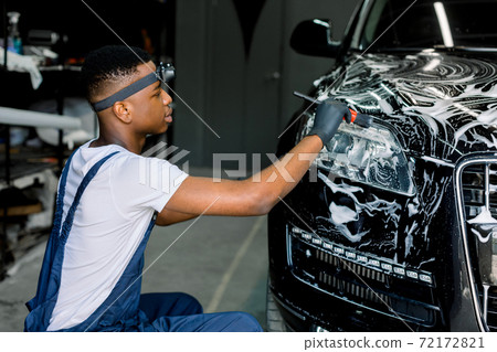 Car detailing wash. Professional car service African male worker in overalls and black rubber gloves, doing detailing wash and cleaning the car with special black brush and cleansing foam Car detailing wash. Professional car service African male worker in overalls and black rubber gloves, doing detailing wash and cleaning the car with special black brush and cleansing foam 72172821
