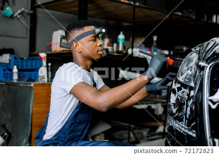 Close up photo of young African worker man in protective gloves, cleaning car headlights with foam and special brush. Carwash and detailing. Washing machine at the station. Manual washing concept. Close up photo of young African worker man in protective gloves, cleaning car headlights with foam and special brush. Carwash and detailing. Washing machine at the station. Manual washing concept. 72172823