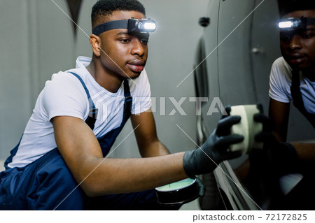 Close up portrait of young handsome African worker of auto repair and detailing service, applying professional solid wax or nanoceramic coating on car body, using the sponge Close up portrait of young handsome African worker of auto repair and detailing service, applying professional solid wax or nanoceramic coating on car body, using the sponge 72172825