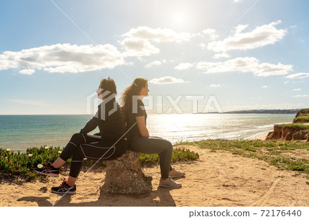 Sport girl and a woman on the shore of the Atlantic Ocean of Portugal, sitting on a stone and talking and thinking about life. Sport girl and a woman on the shore of the Atlantic Ocean of Portugal, sitting on a stone and talking and thinking about life. 72176440