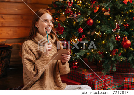 Close-up of ponder smiling young woman wearing winter sweater writing Christmas card with wishes. Close-up of ponder smiling young woman wearing winter sweater writing Christmas card with wishes. 72178171