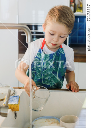 a boy in an apron washes dishes in the kitchen at home 72178337