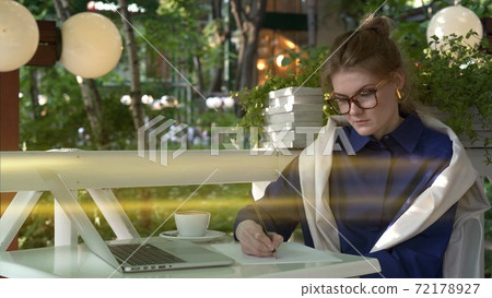 Woman student in eyeglasses with laptop on background of green trees, summer cafe 72178927