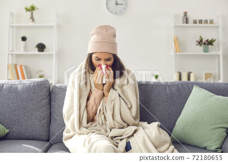Young sick woman in warm clothing, hat sitting on sofa under blanket, blowing nose and feeling ill 72180655