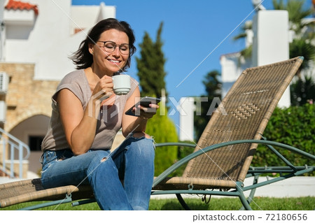 Mature woman relaxing in resort, sitting in chair with cup of coffee and smartphone 72180656
