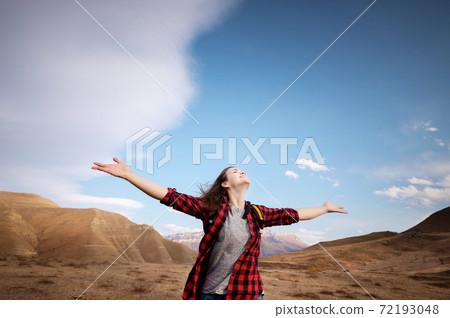 Young happy woman in a T-shirt and jeans stands high in the mountains with her arms outstretched enjoying the view of the mountains and the epic nature of the landscape 72193048