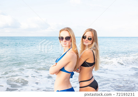 Portrait. Two attractive girls on the beach in bikini look at the camera through sunglasses, against the background of the sea Portrait. Two attractive girls on the beach in bikini look at the camera through sunglasses, against the background of the sea 72193231