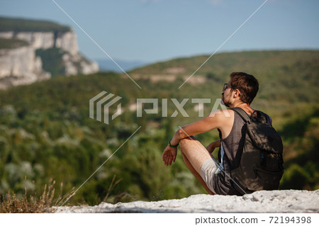 Young male hiker with backpack relaxing on top of a mountain during calm summer sunset Young male hiker with backpack relaxing on top of a mountain during calm summer sunset 72194398
