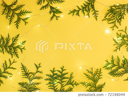 Christmas Or New Year yellow background with frame of green arborvitae branches and luminous Xmas lights on bright yellow with copy space in center. Top view. Horizontal orientation. Christmas Or New Year yellow background with frame of green arborvitae branches and luminous Xmas lights on bright yellow with copy space in center. Top view. Horizontal orientation. 72198049