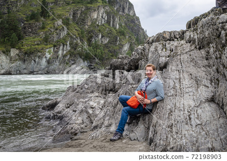 A woman in singlet and jeans with orange backpack near the river against mountains and cloudy sky A woman in singlet and jeans with orange backpack near the river against mountains and cloudy sky 72198903