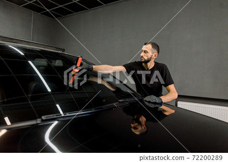 Anti rain coating at auto service. Male worker in black uniform and protective gloves, on a professional car wash service, applying anti rain coating on a windshield of luxury black car Anti rain coating at auto service. Male worker in black uniform and protective gloves, on a professional car wash service, applying anti rain coating on a windshield of luxury black car 72200289