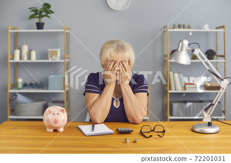 Elderly tired woman sitting with notes, calculator and piggybank and covering stressed face with hands 72201031