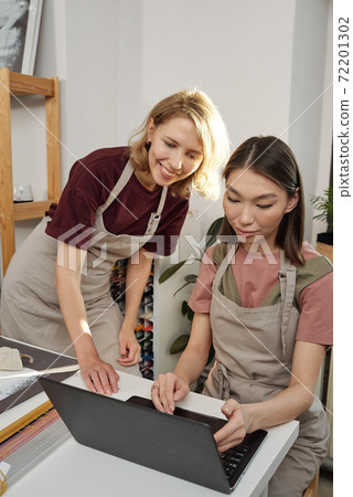 Happy young blond craftswoman in apron bending over colleague using laptop 72201302