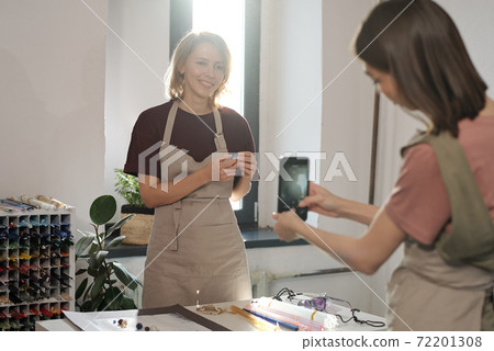 Happy young blond female with handmade glass bead standing by workplace Happy young blond female with handmade glass bead standing by workplace 72201308