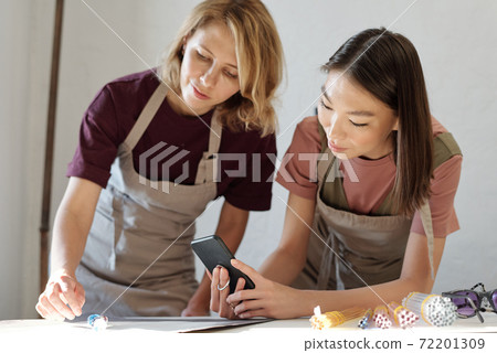 Young woman bending over table and putting glass beads while girl taking photo Young woman bending over table and putting glass beads while girl taking photo 72201309