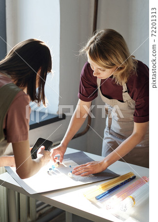Young blond female bending over table and putting glass beads of various shapes Young blond female bending over table and putting glass beads of various shapes 72201313