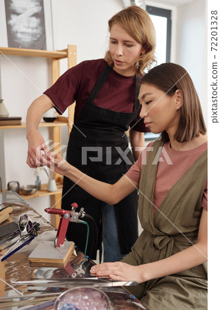 Young female lampworking master in workwear consulting her trainee Young female lampworking master in workwear consulting her trainee 72201328