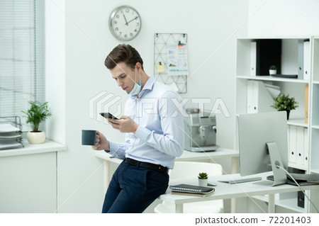 Young businessman in protective mask using smartphone and having tea Young businessman in protective mask using smartphone and having tea 72201403