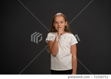 Little girl holding her finger on her lips telling everyone to keep silence wearing white t shirt smiling at the camera isolated on dark grey or black background Little girl holding her finger on her lips telling everyone to keep silence wearing white t shirt smiling at the camera isolated on dark grey or black background 72208752