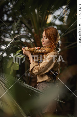 A beautiful girl with natural make-up and red hair stands in the jungle among exotic plants with snake. 72223385