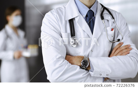 Unknown male doctor in protective mask standing with arms crossed in clinic. Medicine concept during Coronavirus pandemic. Covid 2019 Unknown male doctor in protective mask standing with arms crossed in clinic. Medicine concept during Coronavirus pandemic. Covid 2019 72226376