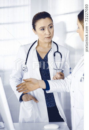 Two female physicians are discussing medical therapy, while standing at the table in a clinic office. Doctors use pc computer at work. Teamwork in medicine 72227080