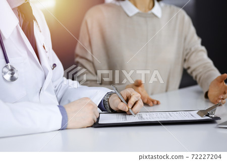 Unknown male doctor and patient woman discussing something while sittingin a darkened clinic and using clipboard, glare of light on the background Unknown male doctor and patient woman discussing something while sittingin a darkened clinic and using clipboard, glare of light on the background 72227204