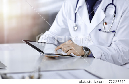 Unknown male doctor sitting and working with tablet computer iin a darkened clinic, glare of light on the background, close-up of hands Unknown male doctor sitting and working with tablet computer iin a darkened clinic, glare of light on the background, close-up of hands 72227211