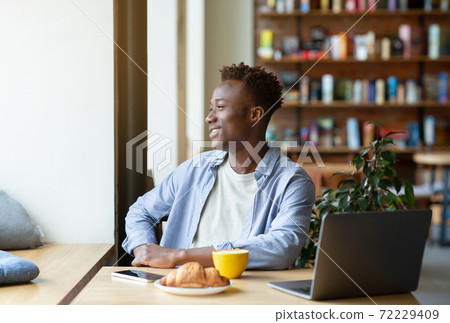 Smiling black guy using laptop computer at cozy cafe, having hot drink with croissant, looking out window 72229409