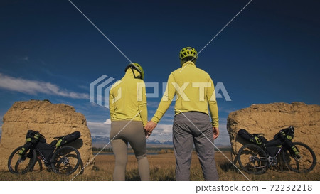 The man and woman travel on mixed terrain cycle touring with bikepacking. The two people journey with bicycle bags. Mountain snow capped, stone arch. 72232218