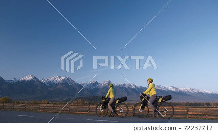The man and woman travel on mixed terrain cycle touring with bikepacking. The couple journey with bicycle bags. Mountain snow capped. 72232712