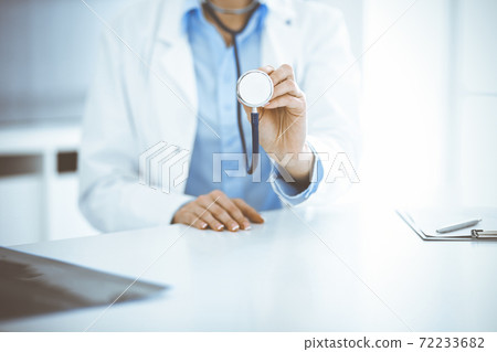 Unknown woman-doctor in blue blouse is holding stethoscope head while sitting at the desk in clinic, close-up. Physician ready to examine and help patient. Medicine concept 72233682