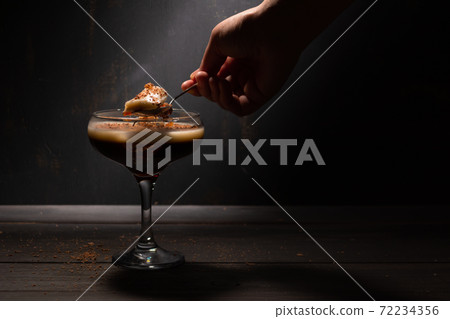 A woman's hand with a spoon takes jelly coffee with milk in a glass bowl, which is sprinkled with chocolate chips. Deep shadows. Rustic. A woman's hand with a spoon takes jelly coffee with milk in a glass bowl, which is sprinkled with chocolate chips. Deep shadows. Rustic. 72234356