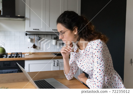 Focused woman wearing glasses looking at laptop screen in kitchen 72235012