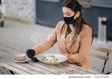 Beautiful young woman eating lunch in cafe outdoors on terrace. Female in black protective mask. Woman take off mask when eating Beautiful young woman eating lunch in cafe outdoors on terrace. Female in black protective mask. Woman take off mask when eating 72237165