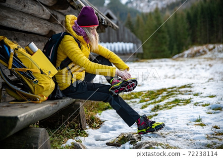 Woman hiker in a yellow down jacket puts yellow crampons on her shoes before going to the mountains. The backpack with equipment is lying next to it. 72238114