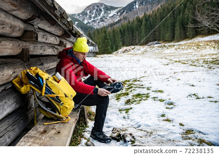 A man tourist in a yellow cap, red down jacket and yellow backpack puts crampons on his shoes. 72238135